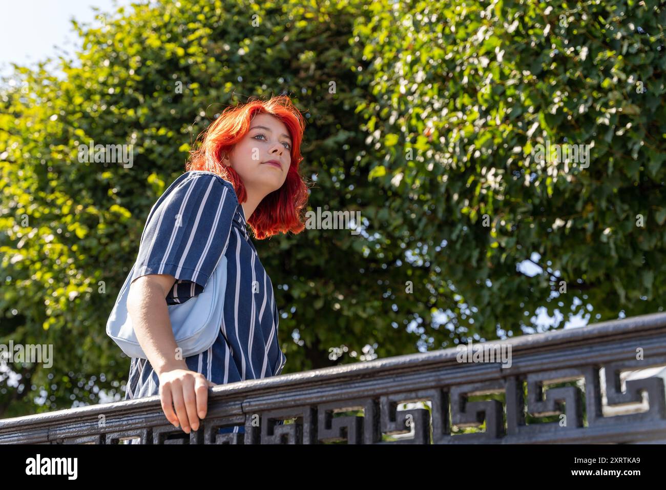 Ein junges Mädchen im Teenageralter mit leuchtend roten Haaren, Teil der Generation Z, macht einen gemütlichen Spaziergang durch einen malerischen Stadtpark, umgeben von üppigem Grün Stockfoto