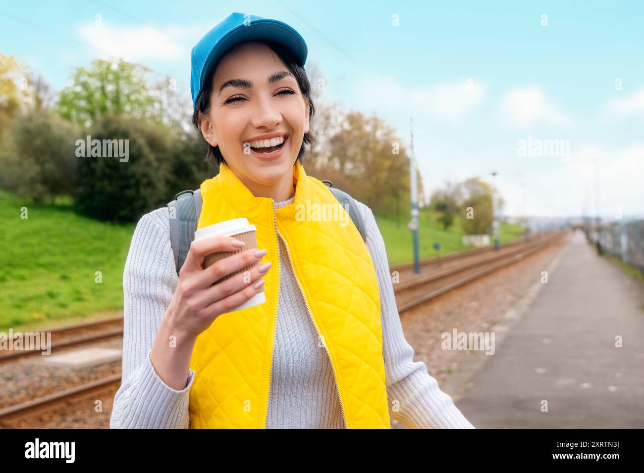 Glückliche junge Frau in gelber Weste und blauer Mütze, die Kaffee zum Mitnehmen, Tee auf der Straße trinkt und Spaß hat. Lifestyle-Konzept Stockfoto