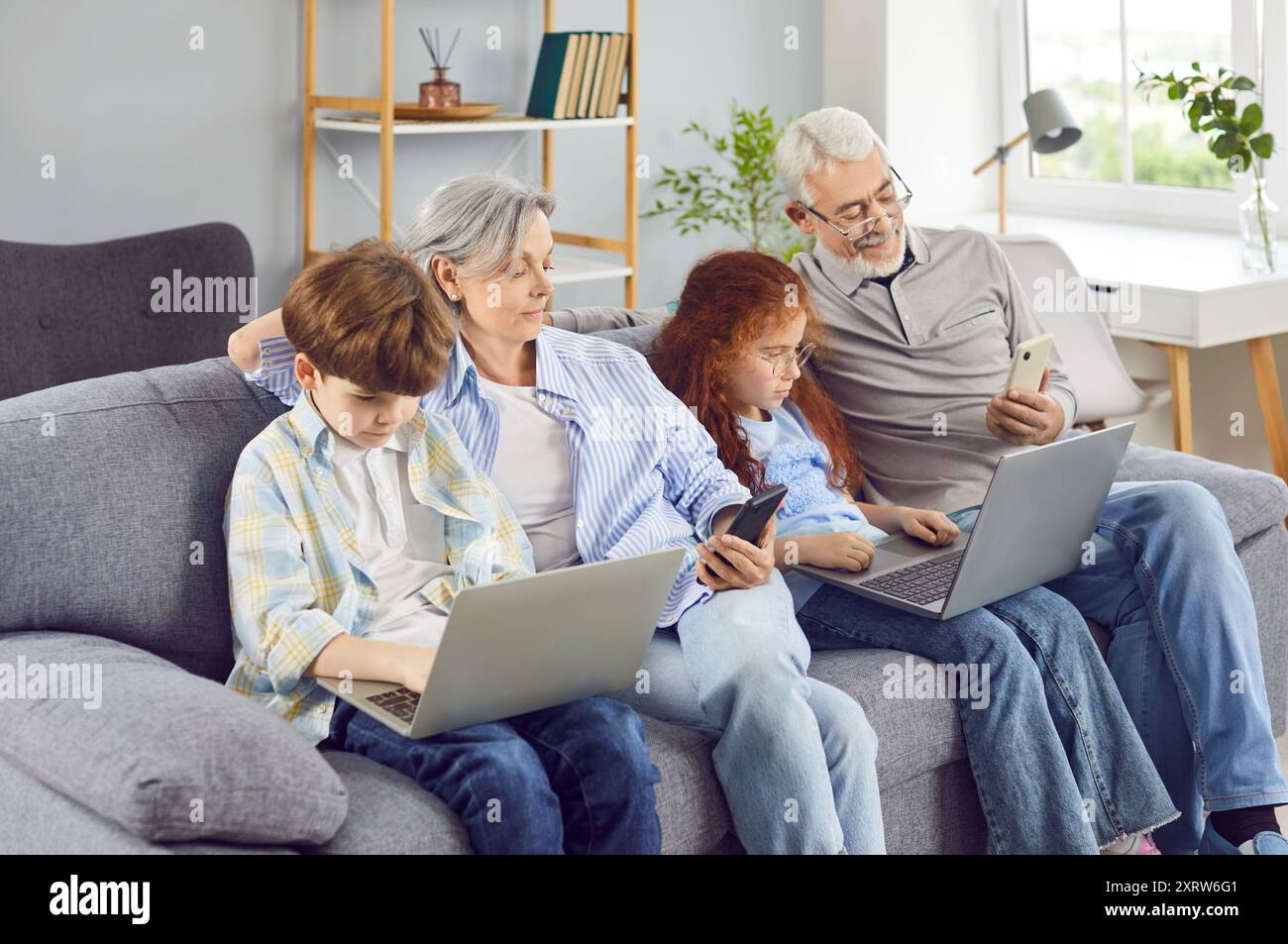 Glückliche Großeltern mit zwei Kindern, die zusammen mit Laptops und Mobiltelefonen auf dem Sofa sitzen. Stockfoto