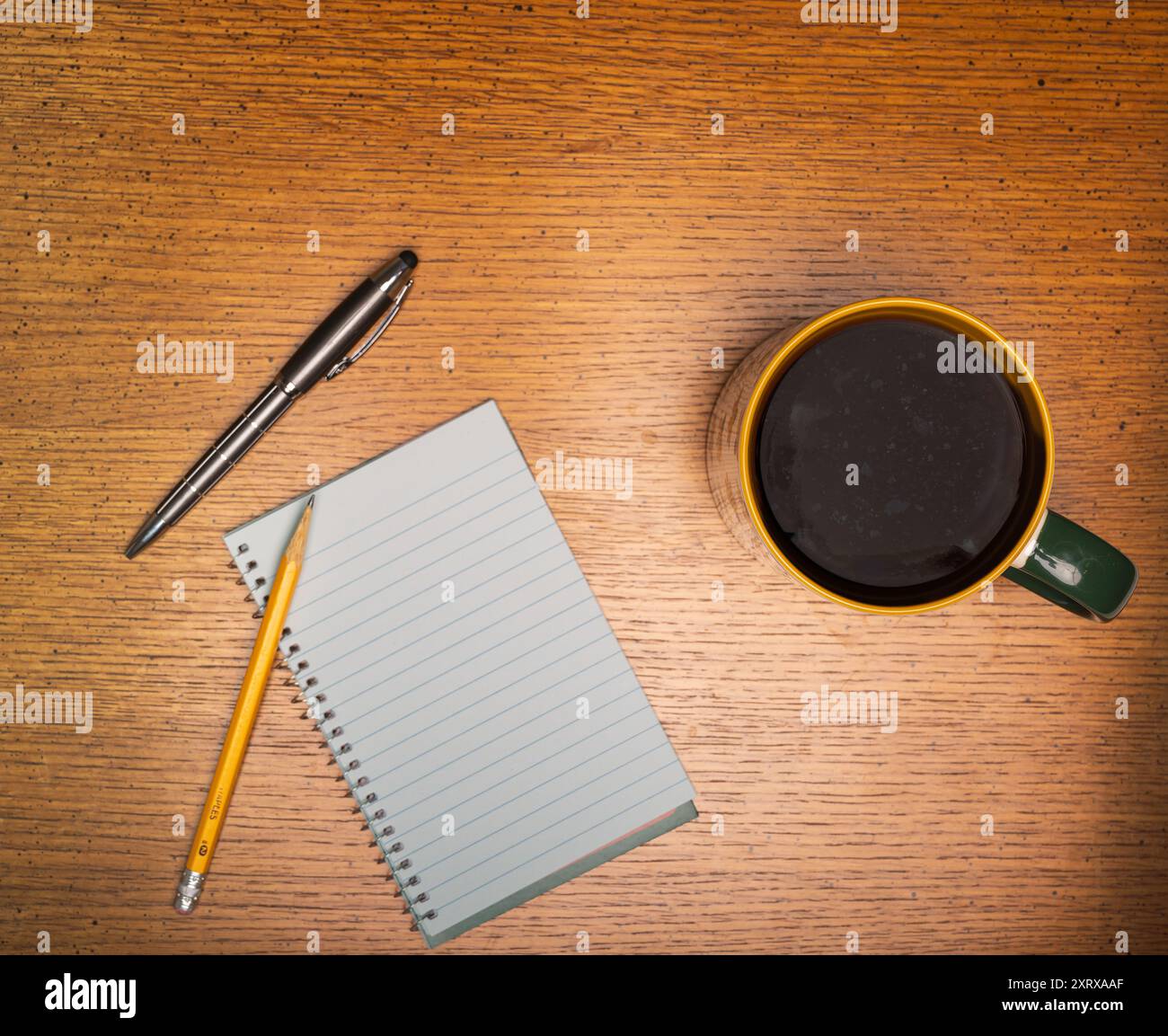 Kaffee in einer goldfarbenen und grünen getrimmten Tasse, die auf einem Tisch aus rauem Holz neben dem spiralförmigen Notizblock und dem Bleistift sitzt Stockfoto