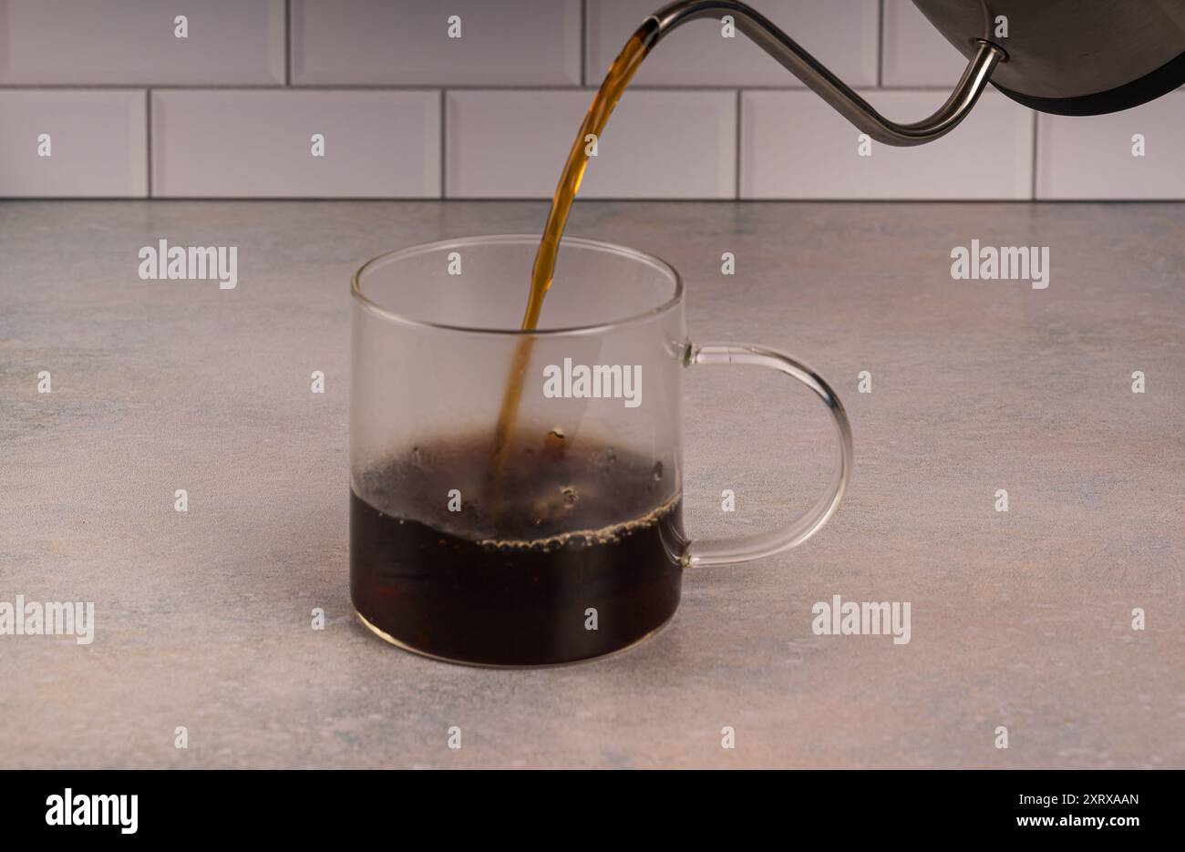 Heißer Kaffee, der in eine Glasschale auf rosa getönter Theke mit weißem U-Bahn-Fliesenhintergrund gegossen wird. Kondenswasser bildet sich an den Seiten des Bechers ab Stockfoto