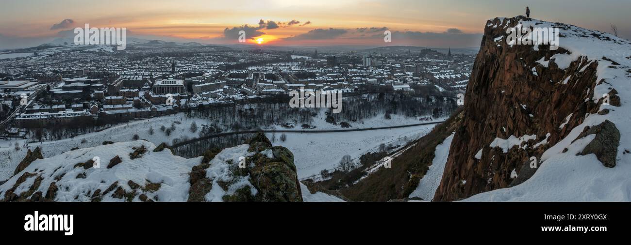 Panoramablick auf Edinburgh, Schottland, mit Schnee vom Arthur Seat, mit Sonnenuntergang hinter der Stadt und Person am Rand der Klippe Stockfoto