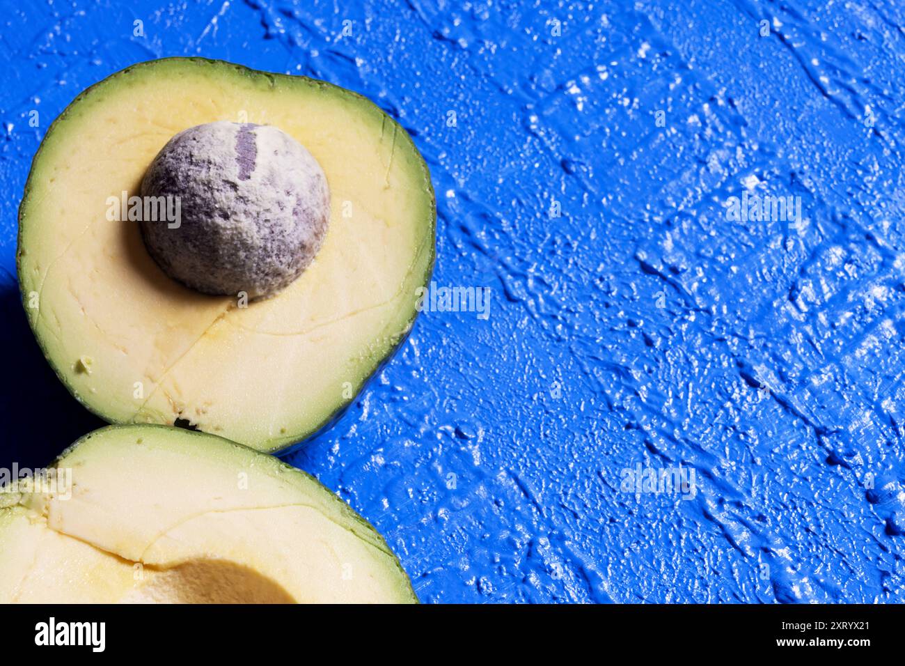 Zwei Teile einer Avocado auf der linken Seite des Bildes, eine Hälfte mit der Grube auf einem strukturierten blauen Hintergrund. Stockfoto