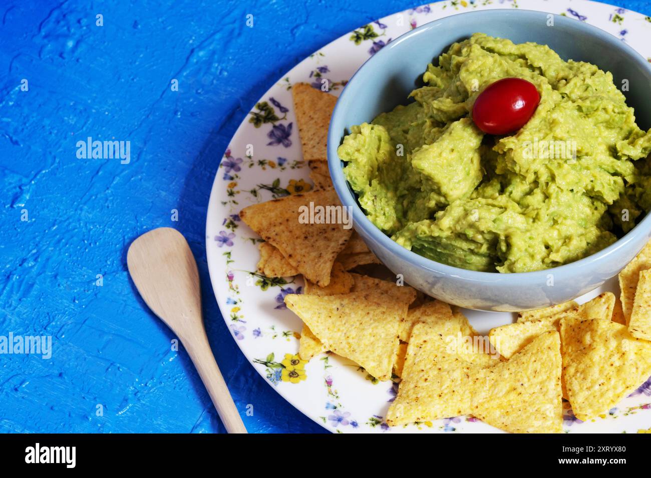 Guacamole in einer blauen Schüssel mit einer Kirschtomate umgeben von Nachos, die auf einem Teller mit Blumen auf blauem Hintergrund dekoriert sind. Stockfoto