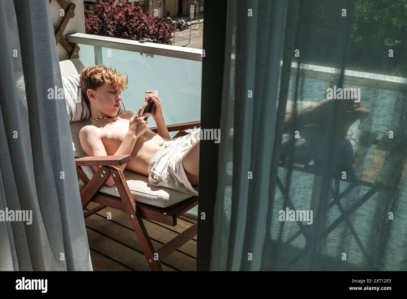Eine junge Person lehnt sich lässig auf einem hölzernen Liegestuhl auf einem sonnigen Balkon, in das sie ein Smartphone einnimmt. Stockfoto