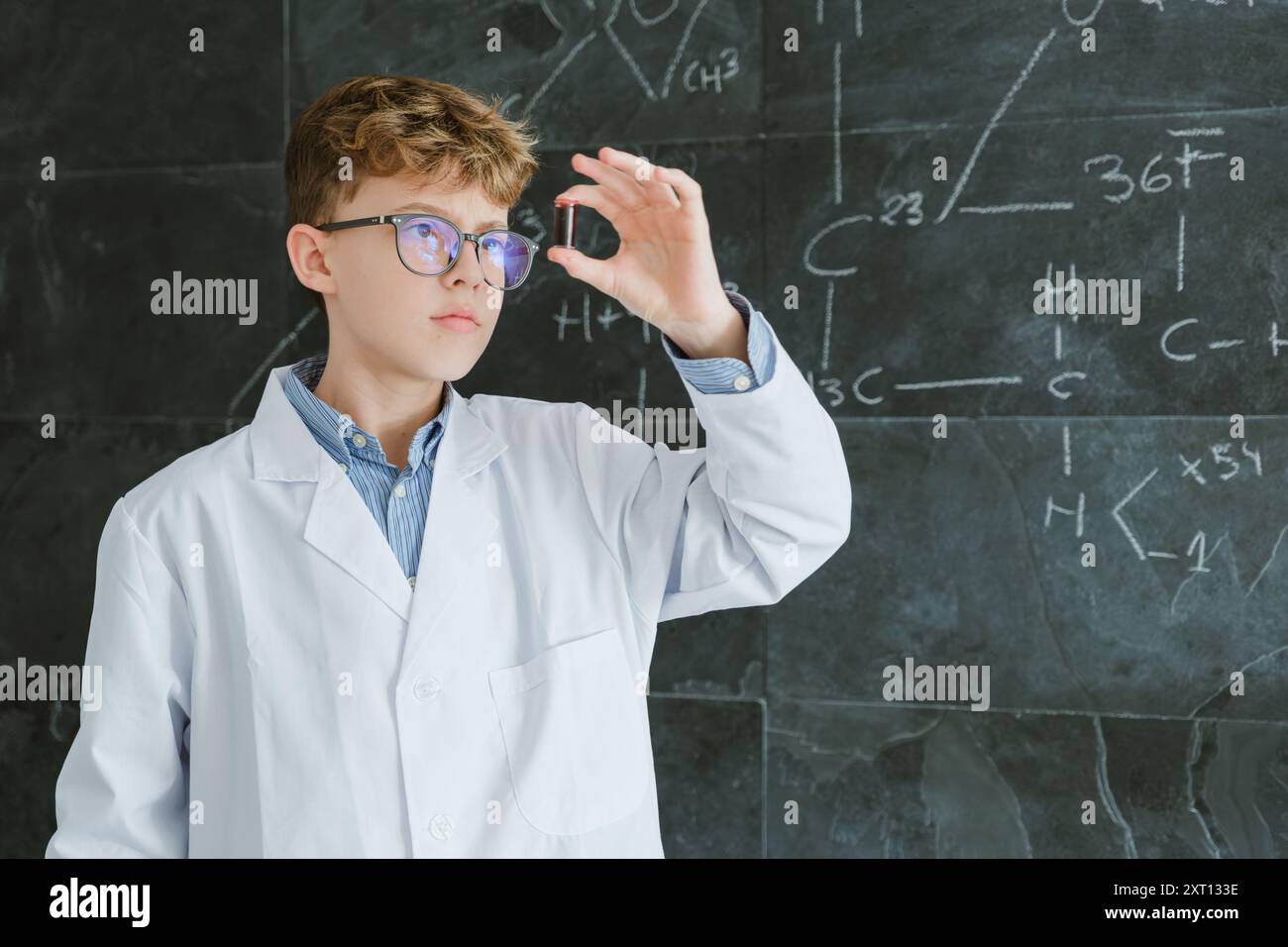 Fokussierter Jugendlicher in Brille, der auf das Reagenzglas blickt, während er vor der Tafel steht, mit schriftlichen chemischen Formeln auf dem Hintergrund im Klassenzimmer Stockfoto
