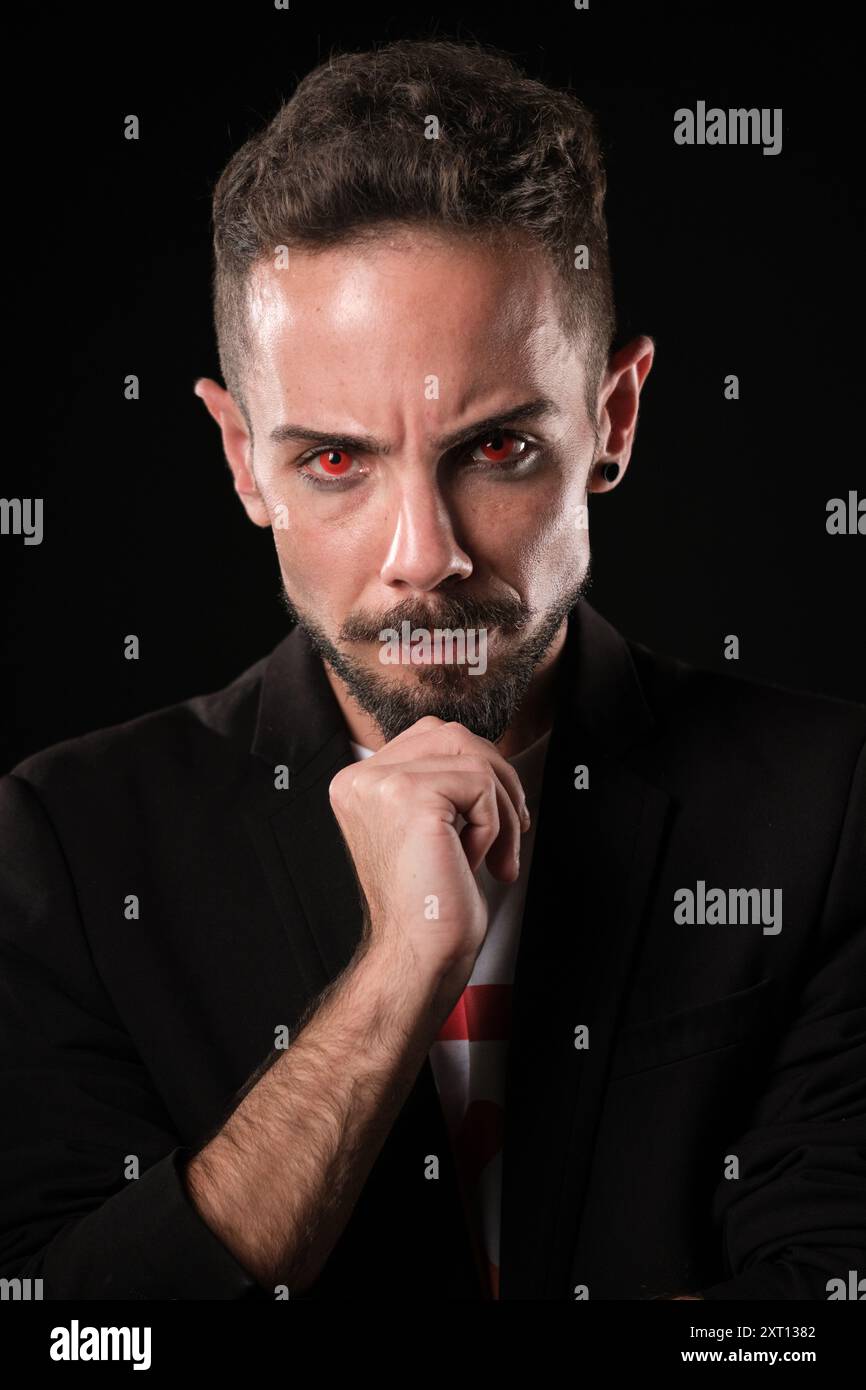 Ein intensiver junger Geschäftsmann in einem schwarzen Anzug starrt selbstbewusst mit einer Hand am Kinn vor dunklem Hintergrund und roten Kontaktlinsen Stockfoto