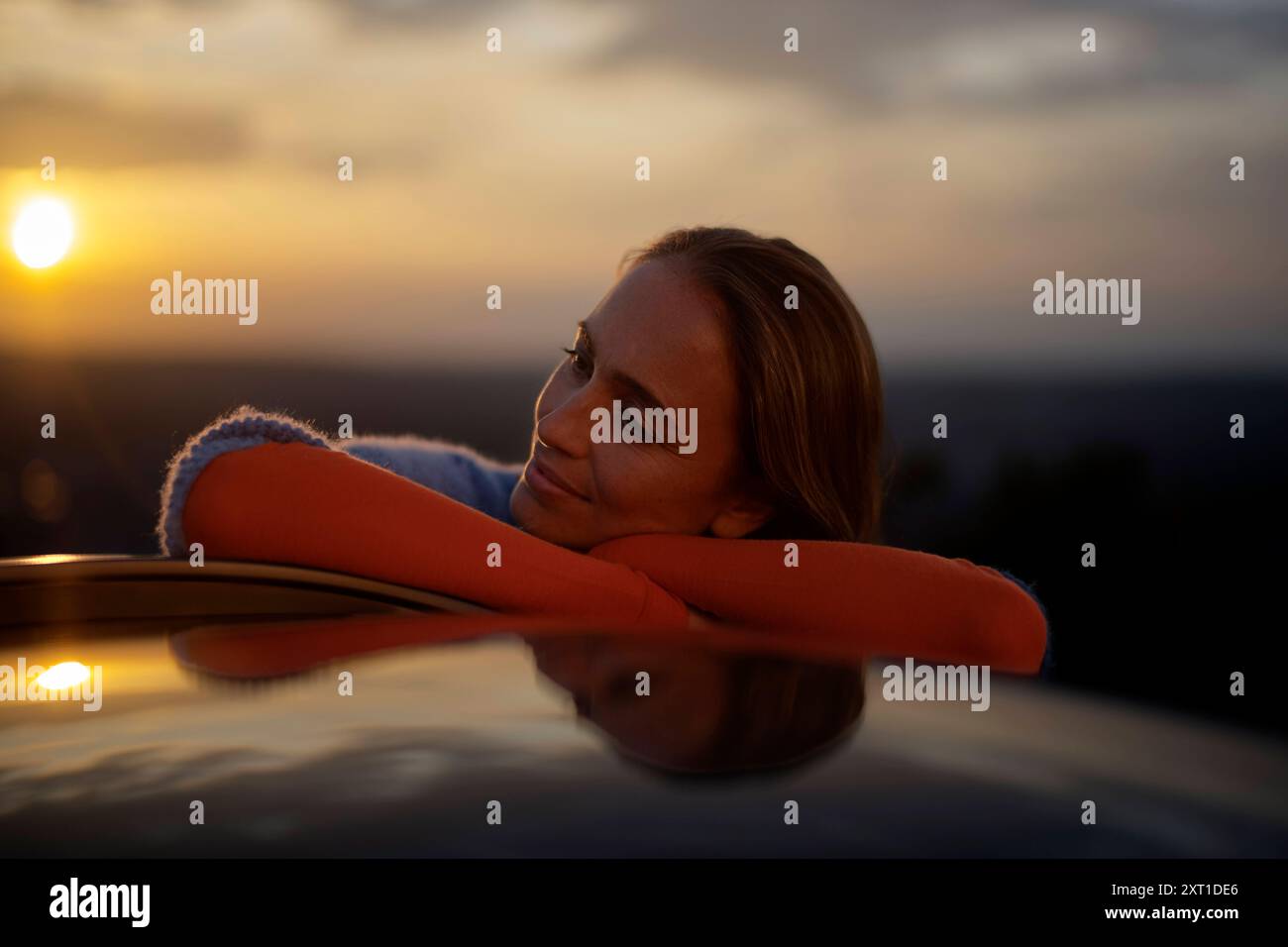 Frau, die sich während des Sonnenuntergangs auf ein Auto lehnt, mit einem zufriedenen Ausdruck. Bola02392 Copyright: XConnectxImagesx Stockfoto