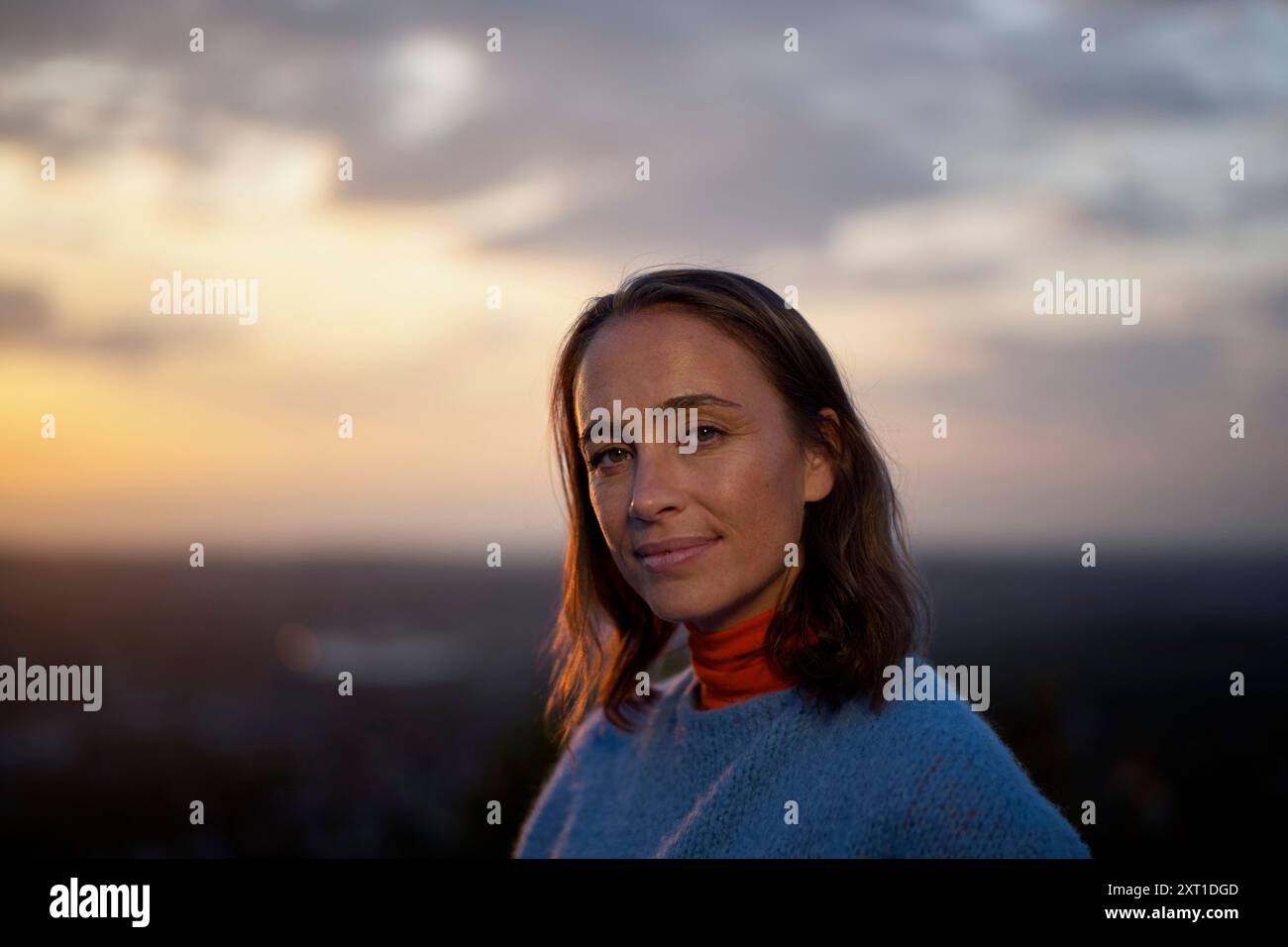 Frau mit einem ruhigen Ausdruck posiert bei Sonnenuntergang mit einem sanft verschwommenen Stadtbild Hintergrund. Bola02412 Copyright: XConnectxImagesx Stockfoto