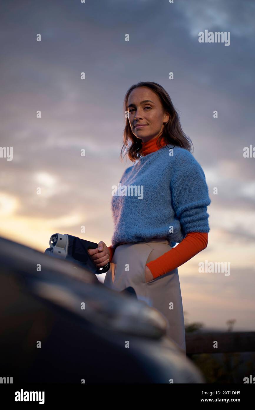Frau, die bei Sonnenuntergang mit einem besinnlichen Ausdruck an einem Auto steht. Bola02410 Copyright: XConnectxImagesx Stockfoto
