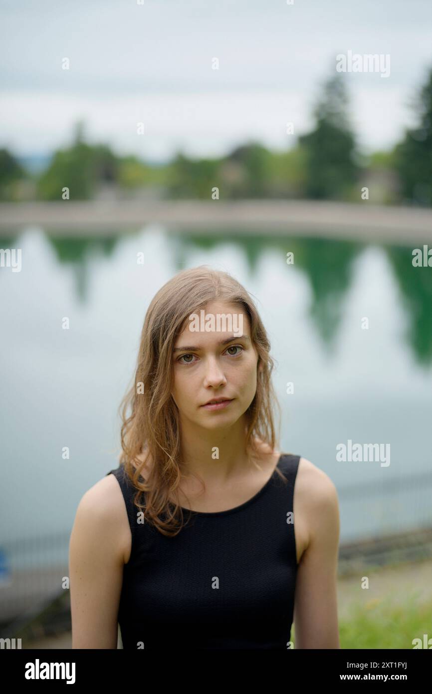Eine junge Frau mit einem feierlichen Ausdruck, die vor einem ruhigen See steht, mit Spiegelungen von Bäumen im Wasser. Bola02504 Copyright: XConnectxImagesx Stockfoto