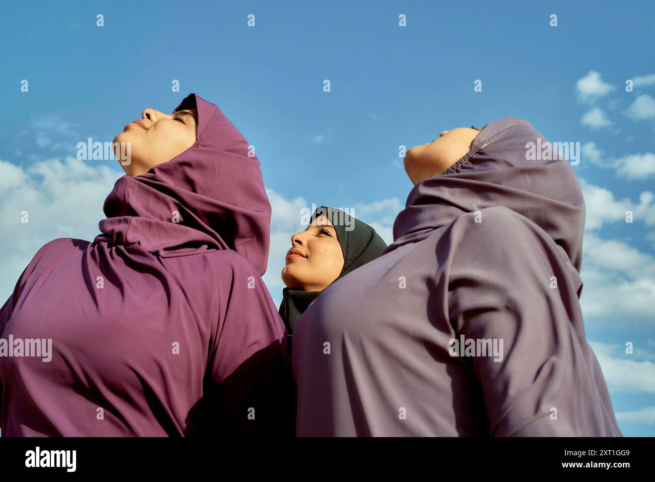 Drei Frauen in violettem Hijabs stehen vor einem blauen Himmel und blicken mit heiterem Ausdruck nach oben. Bola02592 Copyright: XConnectxImagesx Stockfoto