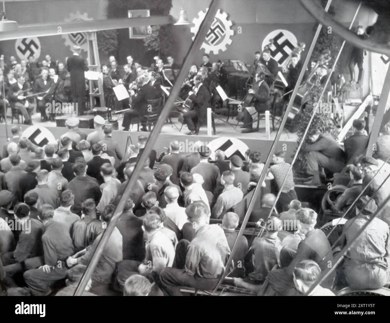 Ein Foto zeigt Arbeiter, die sich zu einer musikalischen Feier in einer Stahlfabrik versammelten und die Bemühungen des NS-Regimes widerspiegeln, die kulturelle Einheit und Moral unter den Arbeitskräften zu fördern. Diese Ereignisse waren Teil der breiteren Propagandakampagne, um Industriearbeiter als integraler Bestandteil der Stärke und des Erfolgs der Nation darzustellen. Stockfoto