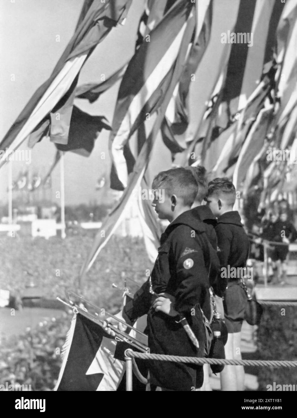 Ein Foto zeigt Jungen der Hitlerjugend, die Instrumente auf der Spitze eines Stadions spielen. Die 1926 gegründete Hitlerjugend war ein zentraler Bestandteil der Indoktrinierungsbemühungen Deutschlands, die sich auf körperliche Fitness, militärisches Training und Loyalität gegenüber Adolf Hitler konzentrierte. Ereignisse wie diese, mit Fanfaren, Fahnen und Liedern der Freiheit, sollten Einheit und Nationalstolz fördern und die Jugend darauf vorbereiten, dem Dritten Reich zu dienen. Stockfoto