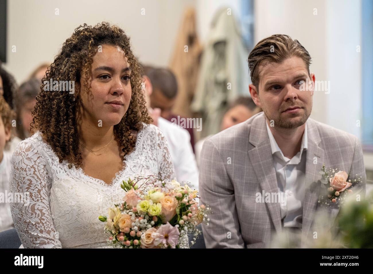 Eine Braut in einem Spitzenkleid mit einem Blumenstrauß sitzt neben dem Bräutigam in einem hellgrauen Anzug, beide sind aufmerksam bei einer formellen Veranstaltung. Panc05783 Copyright: Stockfoto