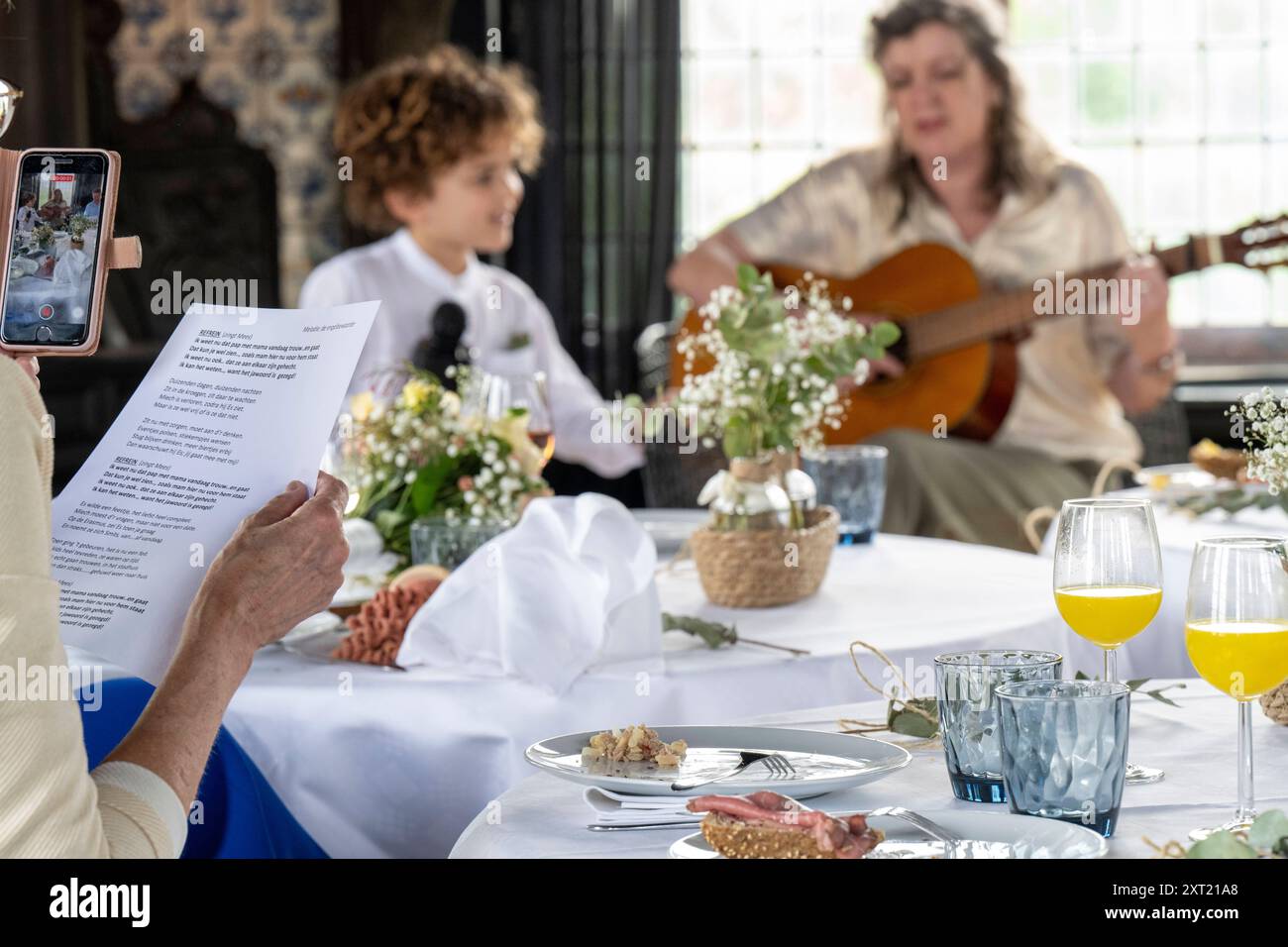 Eine Familie genießt eine feierliche Mahlzeit zusammen mit Live-Gitarrenmusik in einem warm beleuchteten Raum, in dem Speisen, Getränke und ein Smartphone den Moment festhalten Stockfoto
