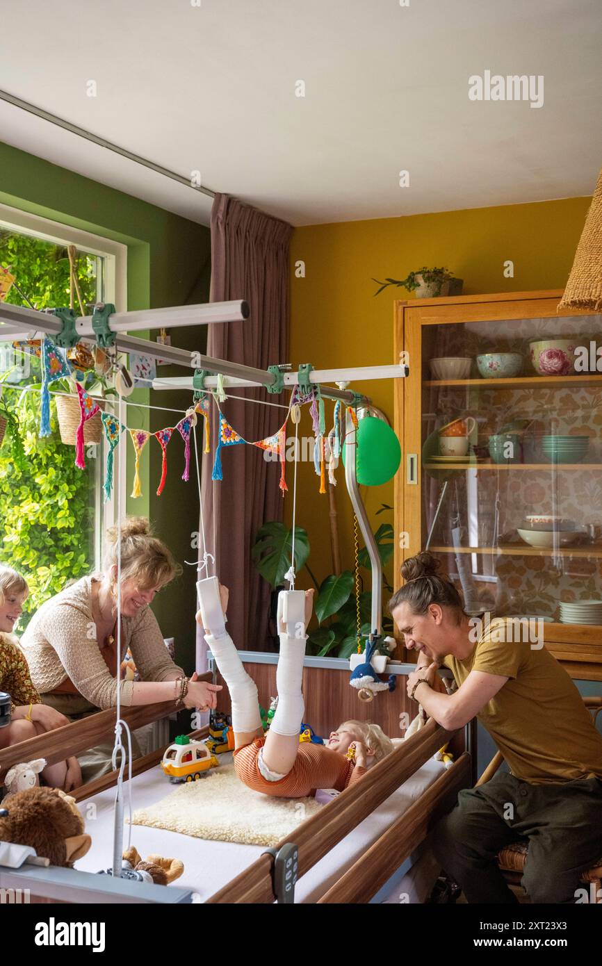 Ein Kind liegt auf dem Rücken und spielt mit bandagierten Beinen in der Luft, während ein Erwachsener unter Spielzeugen zusieht. Familie um sie zu unterstützen Panc06107 Copyright: Stockfoto