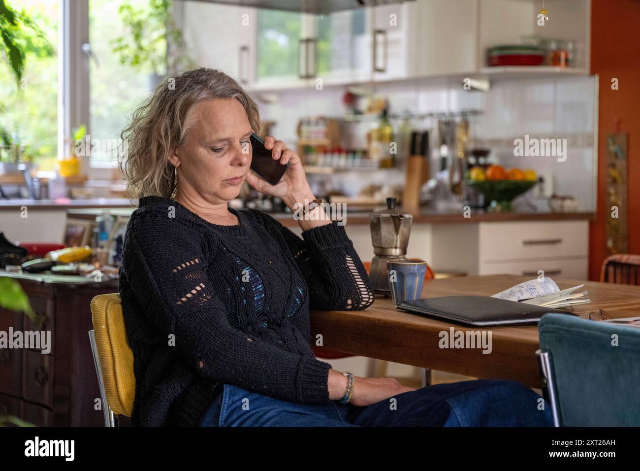 Eine erwachsene Frau sitzt an einem Esstisch und redet mit ihrem Smartphone und sieht besorgt aus, wie ein offenes Notebook vor ihr liegt. Panc06316 Copyright: XConnec Stockfoto