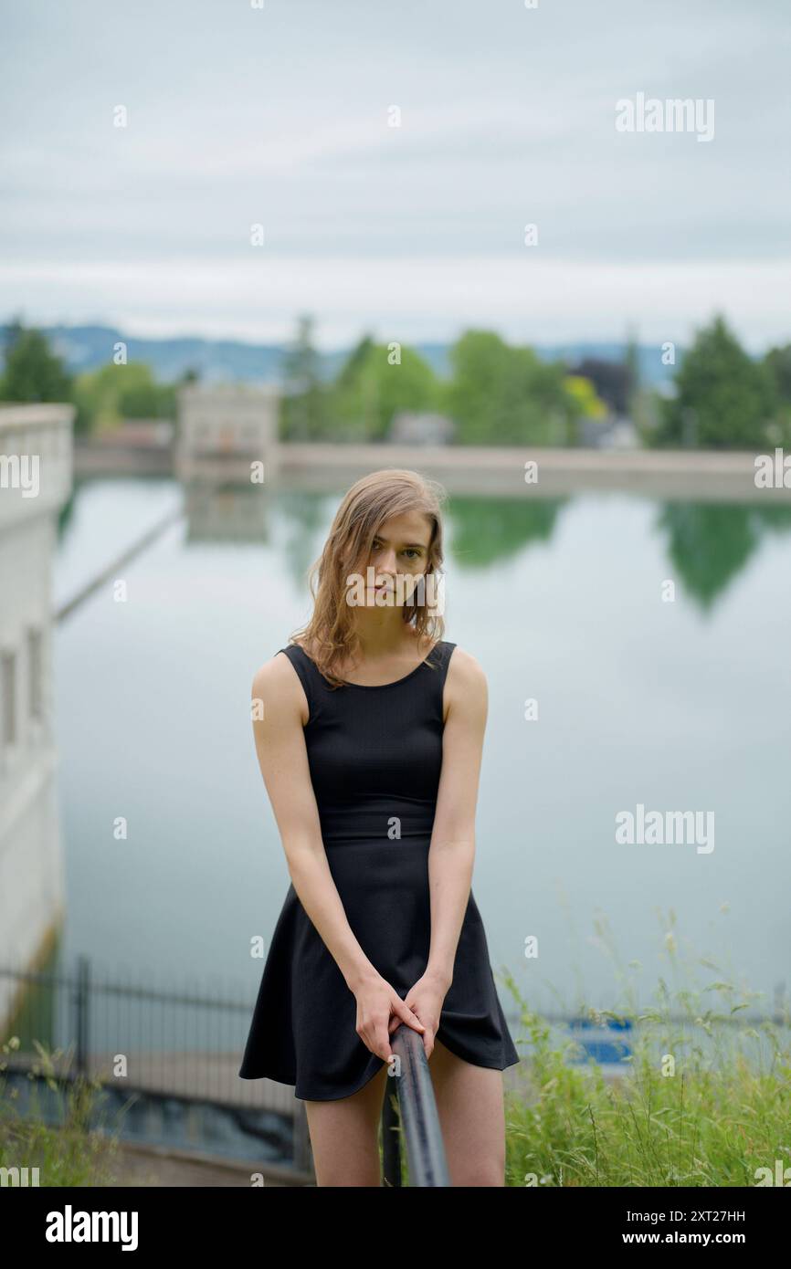 Eine junge Frau in einem schwarzen Kleid steht an einem Geländer mit einem ruhigen See und Grün im Hintergrund. Bola02771 Copyright: XConnectxImagesx Stockfoto