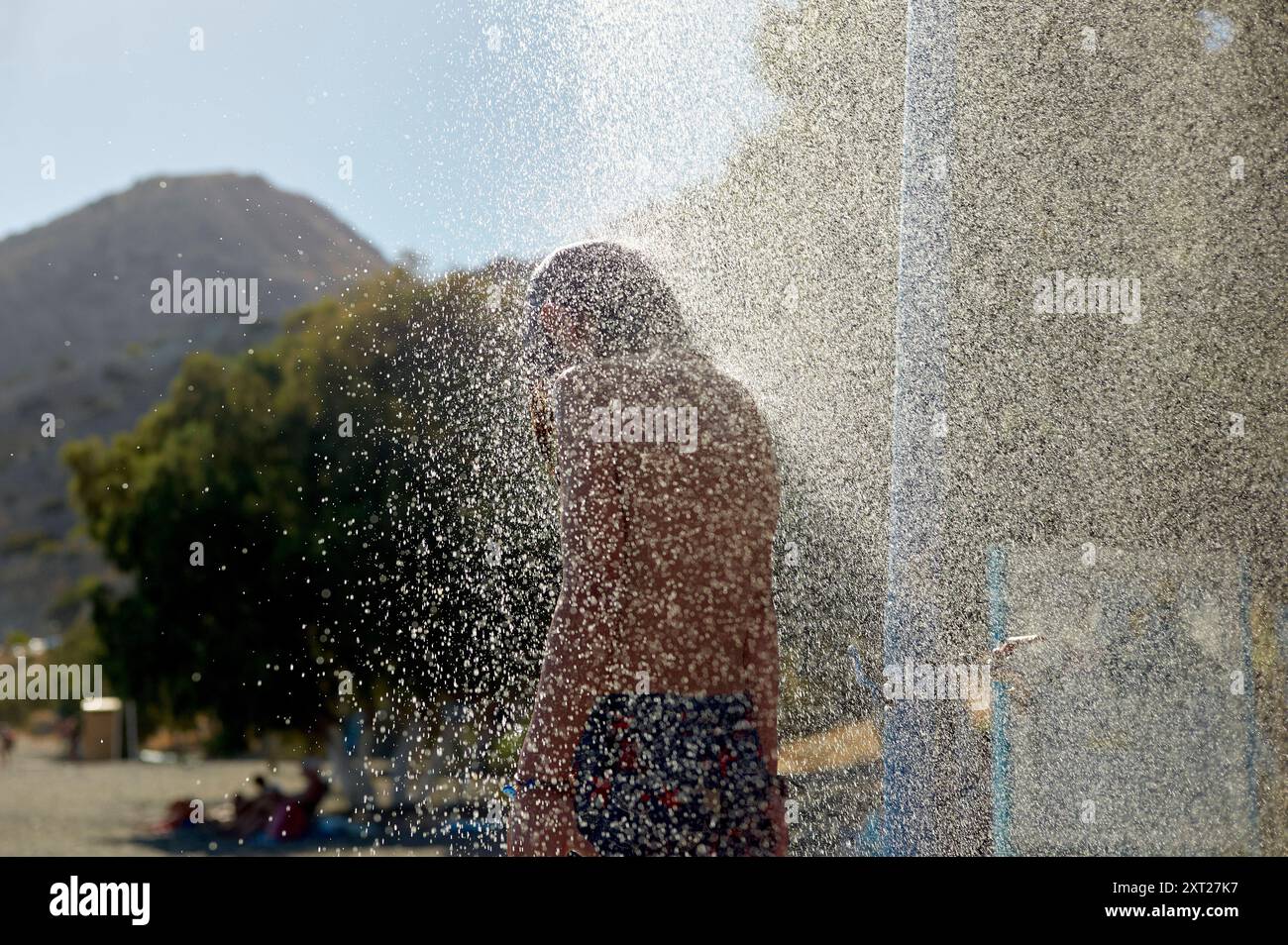 Junge, der draußen duscht, mit Wassertropfen in der Luft, die das Sonnenlicht einfangen. Bola02777 Copyright: XConnectxImagesx Stockfoto