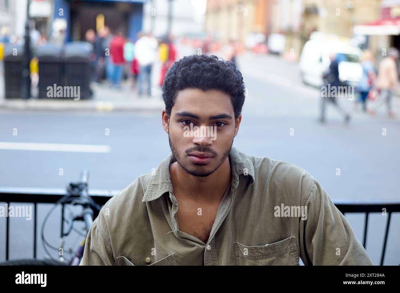 Ein junger Mann mit lockigen Haaren schaut ruhig in die Kamera, mit einer verschwommenen Straßenszene im Hintergrund. Bola02865 Copyright: XConnectxImagesx Stockfoto
