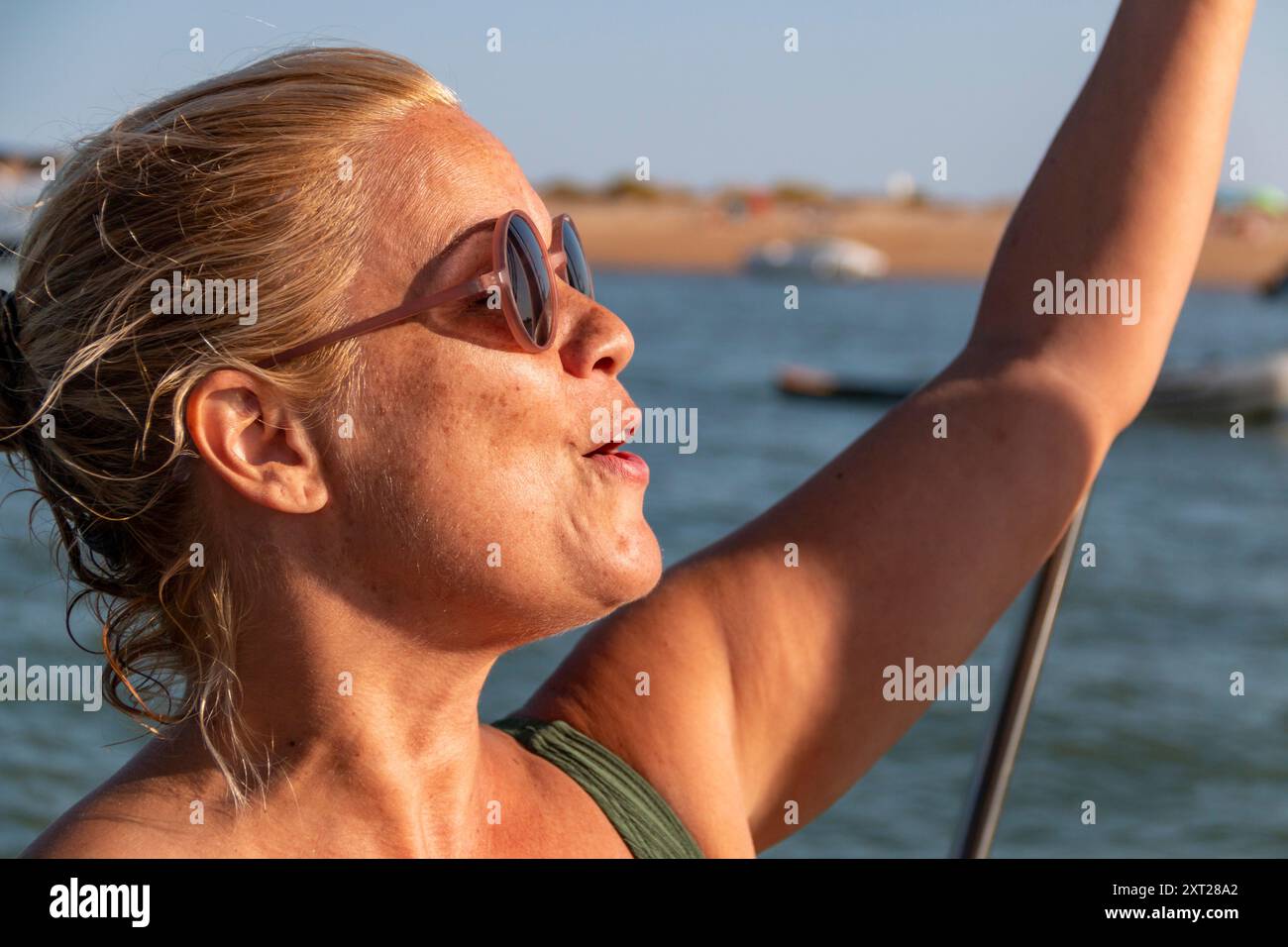 Blonde Frau mit lockeren Haaren und Sonnenbrille beugt sich über die Seite eines Bootes über die Wellen und das blaue Meer Stockfoto