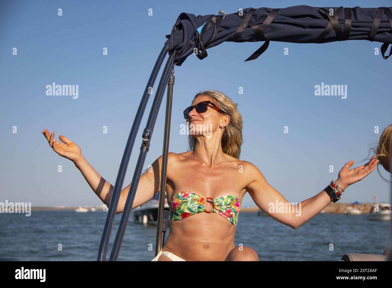 Blonde Frau mit lockeren Haaren und Sonnenbrille beugt sich über die Seite eines Bootes über die Wellen und das blaue Meer Stockfoto