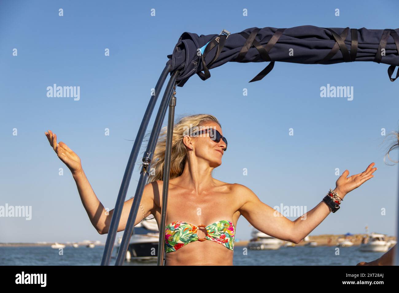 Blonde Frau mit lockeren Haaren und Sonnenbrille beugt sich über die Seite eines Bootes über die Wellen und das blaue Meer Stockfoto