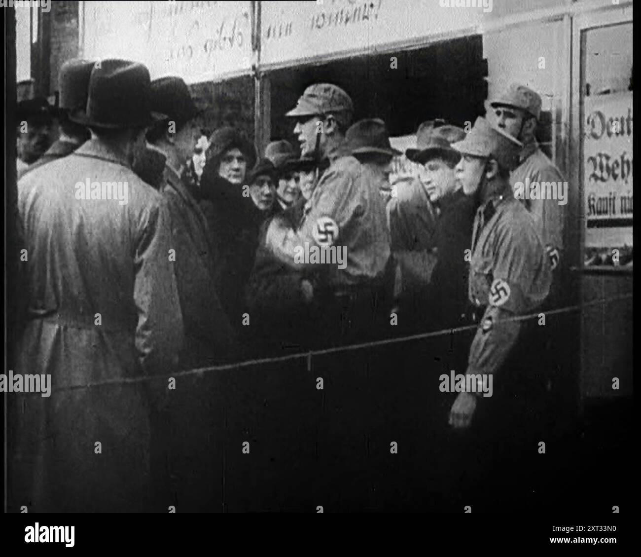 Männer in Nazi-Uniformen und Zivilisten stehen vor einem Geschäft mit antijüdischen Plakaten, 1933. "Wieder einmal, in der langen Geschichte der Welt, Qualen für die Juden". Aus „Time to Remember – The Time of the Monster“, 1933 (Reel 4); ein Dokumentarfilm über die Ereignisse von 1933, Rise of Roosevelt und Hitler. Stockfoto