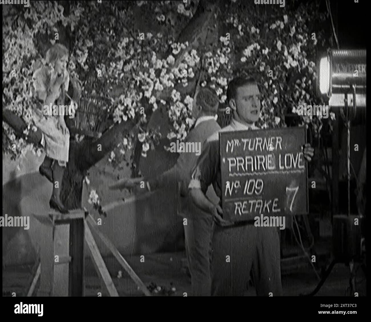 Weibliche Schauspielerin sitzt in einem Blossom Tree, während ein männlicher Zivilist mit ihr spricht. Ein Filmtechniker hält ein Clapper Board mit der Aufschrift „Mr. Turner Prairie Love No. 109 Rake 7“, 1920er Elstree, im Großen und Ganzen das Hollywood Großbritanniens in den Zwanzigern, aber ohne Palmen und Sonne. Aber auf den Bühnen mit Zinndach konnte man es zu jeder Jahreszeit schaffen. Aus „Time to Remember – Come the Dawn“, 1925 (Rolle 1); werfen Sie einen Blick auf die britische Filmindustrie der 1920er Jahre – Clips aus großen Stummfilmen und Wochenschauen. Stockfoto