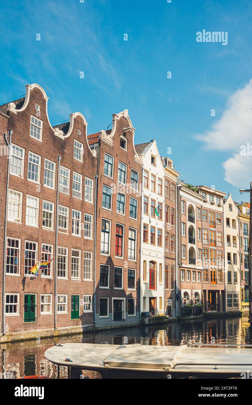 Die helle Sonne beleuchtet klassische Amsterdamer Grachtenhäuser mit charakteristischen Giebelfassaden entlang einer ruhigen Wasserstraße und fängt das Wesen der niederländischen Architektur ein Stockfoto
