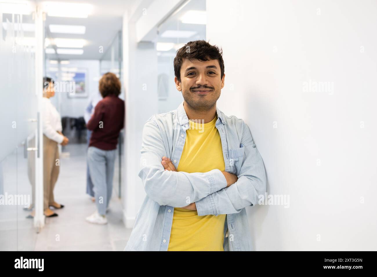 Ein junger Mann lächelt selbstbewusst in die Kamera, die Arme überkreuzt, in einem gut beleuchteten Korridor der englischen Akademie, während zwei Kollegen im Hintergrund diskutieren Stockfoto