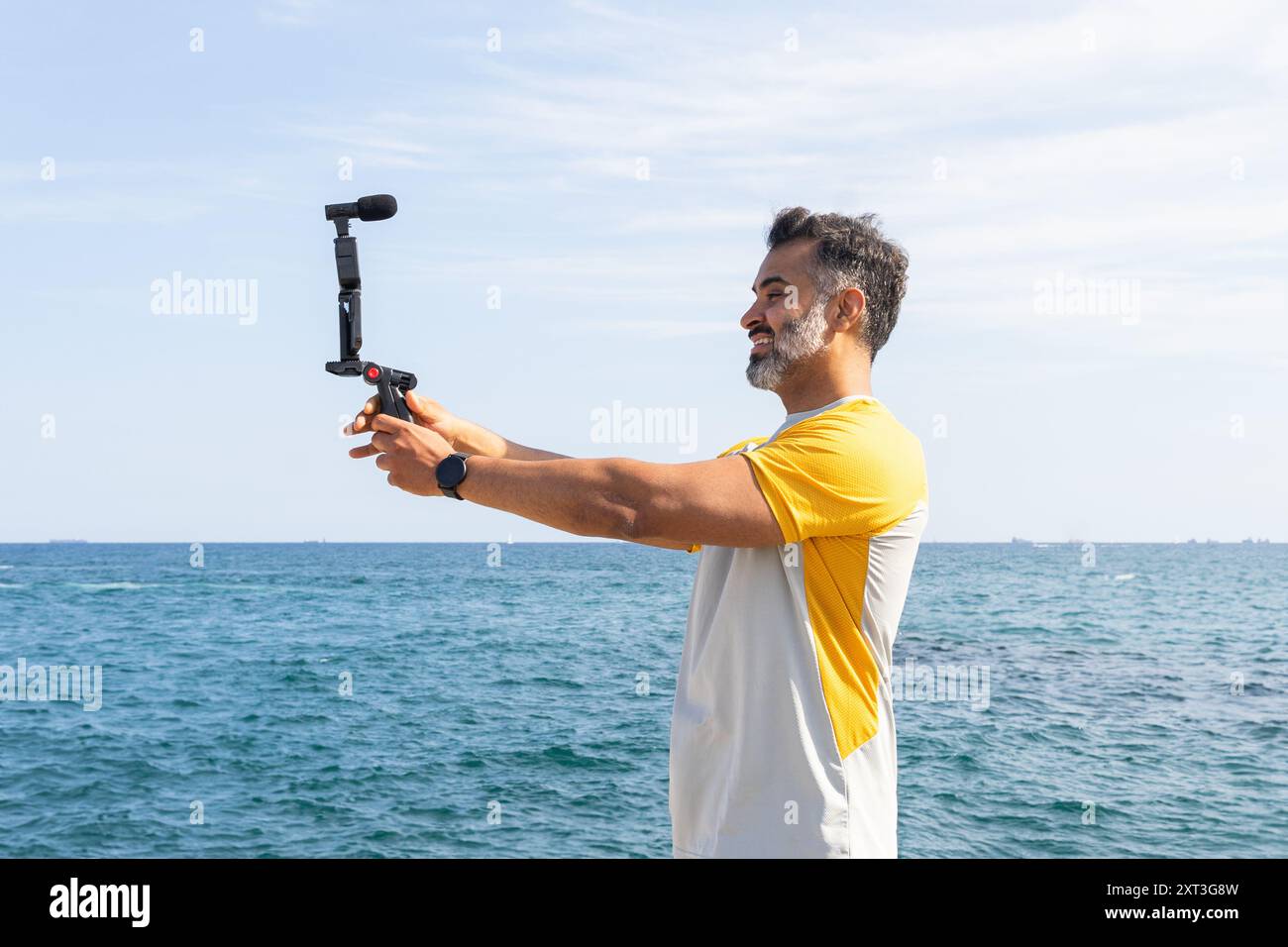 Ein lächelnder indischer Mann in einem sportlichen Outfit nimmt ein Gimbal auf, um ein Video am Meer aufzunehmen und auf den Bildschirm des Geräts zu schauen. Stockfoto