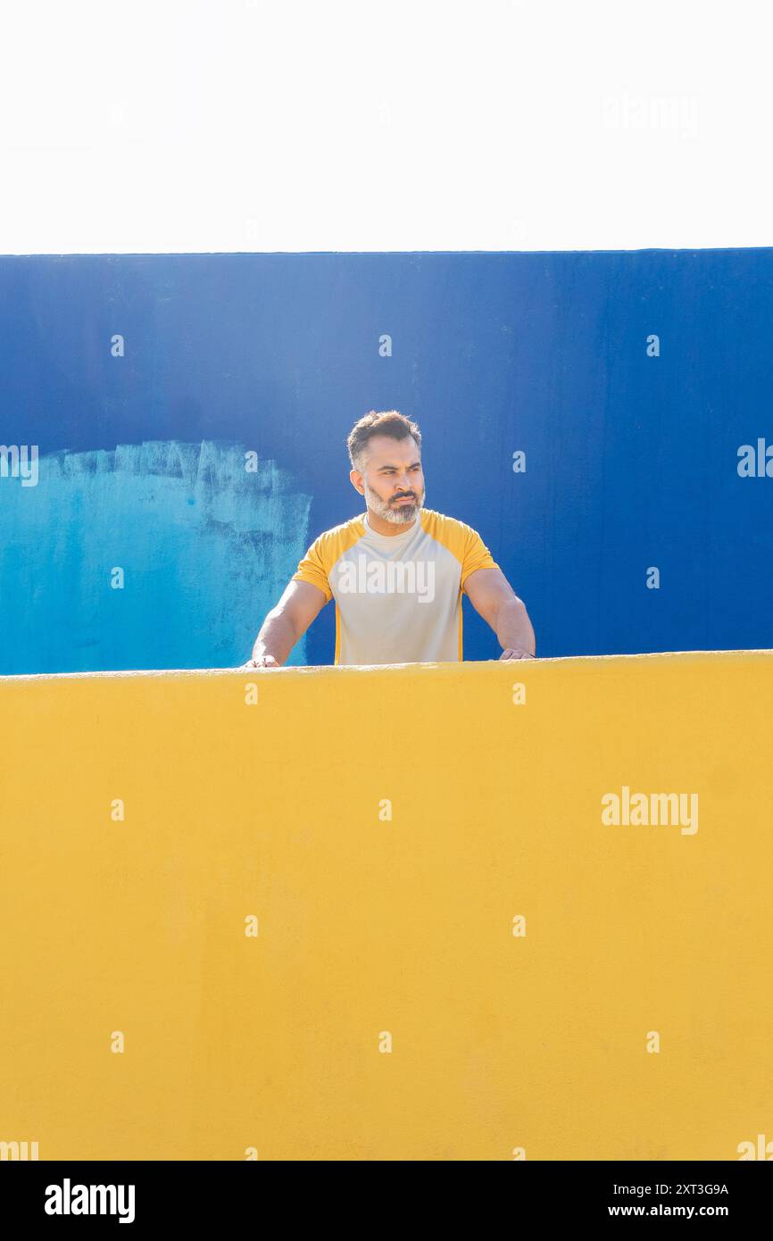 Ein indischer Mann in Gelb-weiß-Sportbekleidung liegt an einer gelben Wand vor einer kontrastierenden blauen Wand und blickt an einem Sommertag von der Kamera weg. Stockfoto