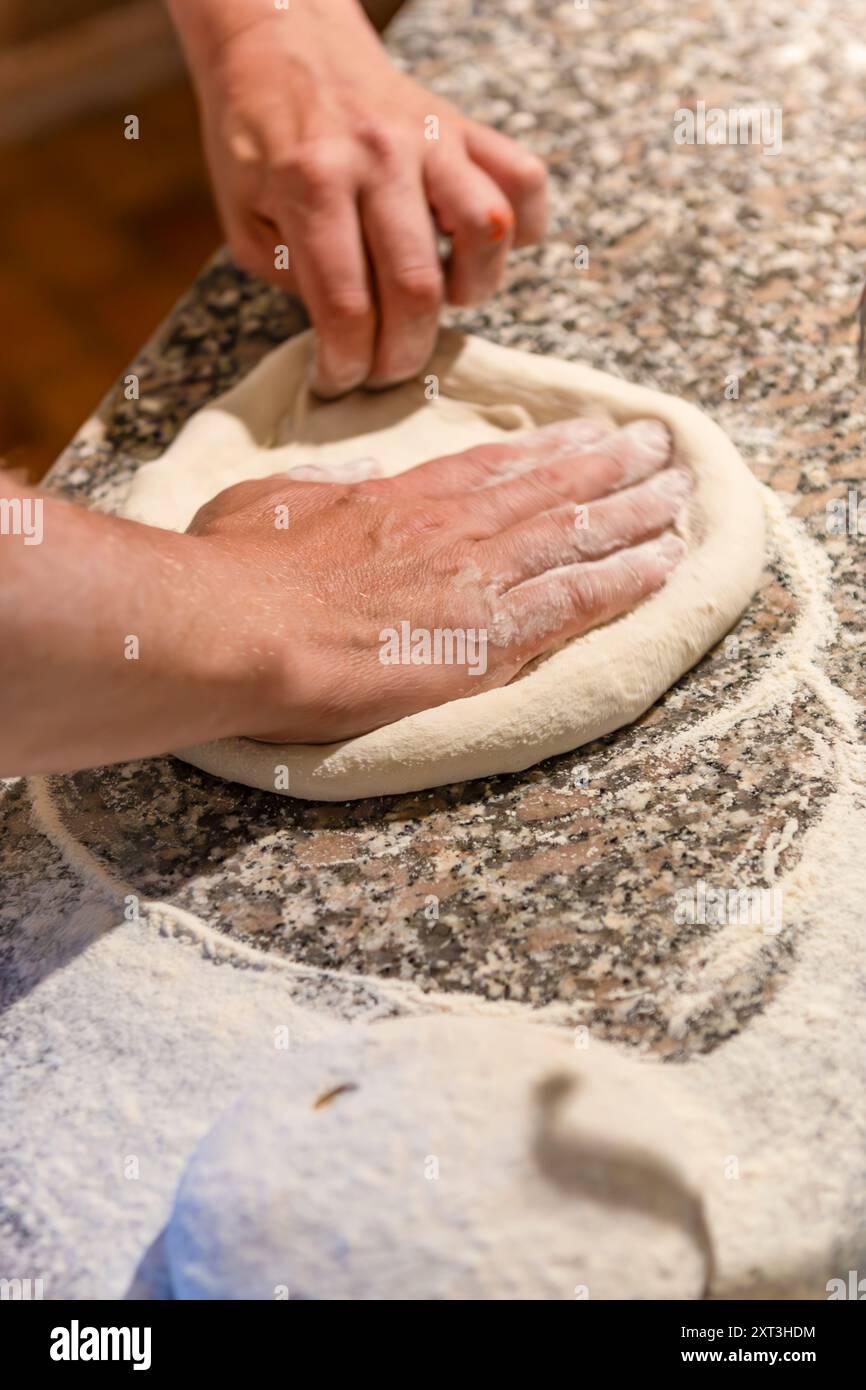 Nahaufnahme der Hände, die frischen Pizzateig auf einer Küchenarbeitsfläche aus Granit Formen, die mit Mehl verstreut ist, um den Prozess der Pizzaherstellung hervorzuheben Stockfoto