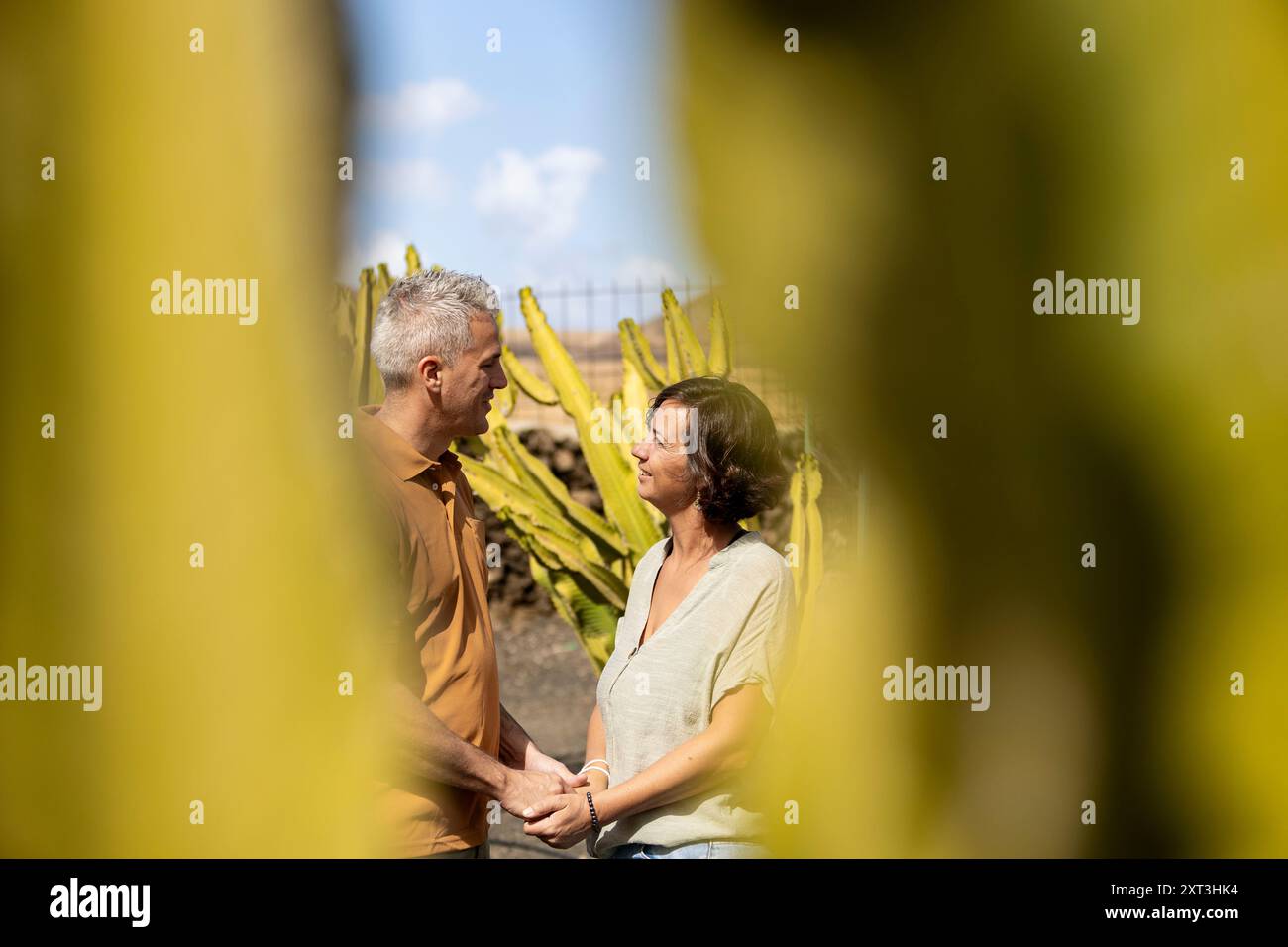 Ein reifer Mann und eine reife Frau stehen nahe, halten die Hände und blicken einander liebevoll an. Verschwommenes Laub umrahmt sie und hebt ihre tiefe Verbindung hervor Stockfoto