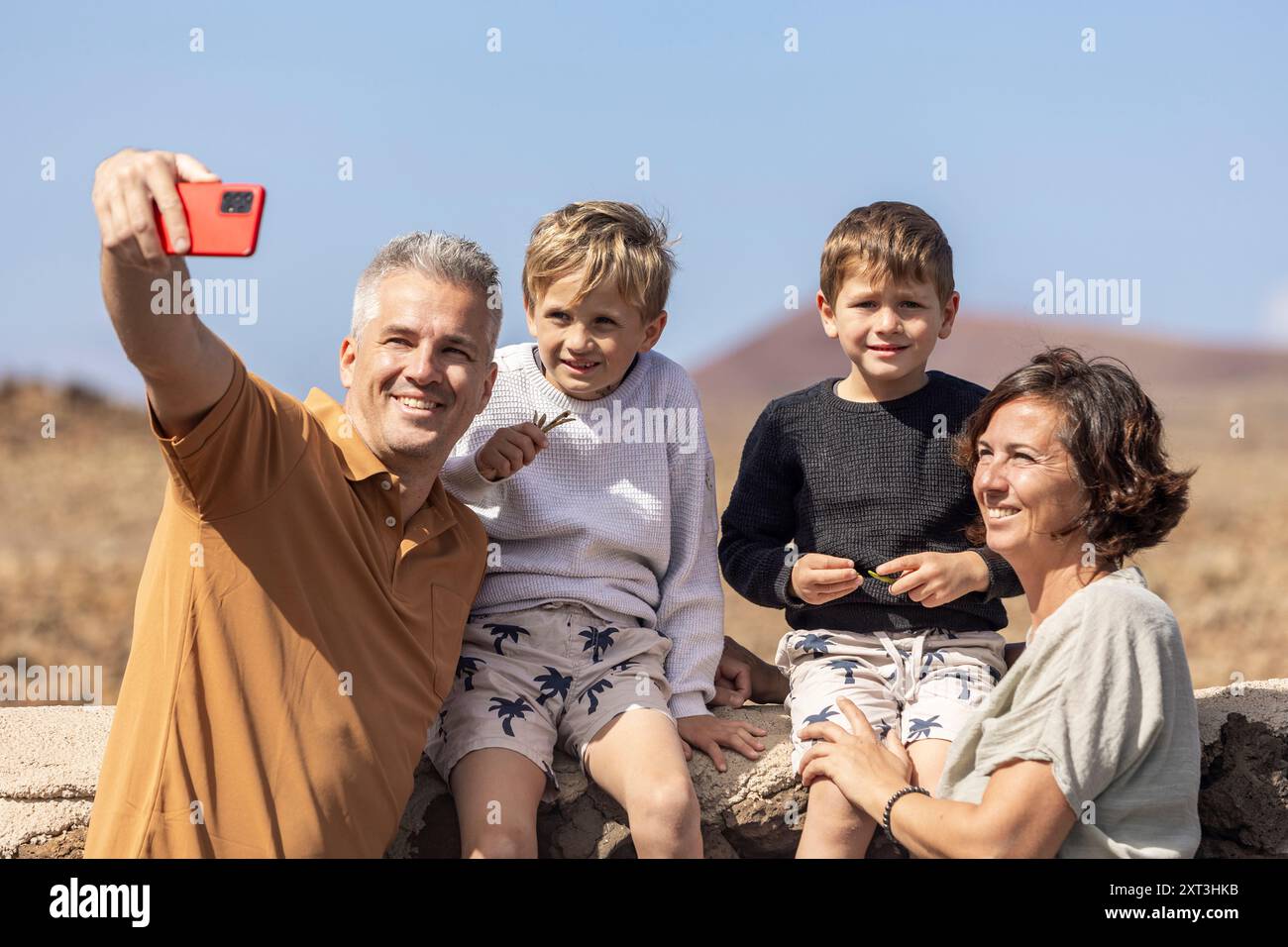 Eine vierköpfige Familie mit Mutter, Vater und ihren beiden Jungs genießt einen sonnigen Tag im Freien. Der Vater macht ein Selfie mit einem Smartphone, Captu Stockfoto