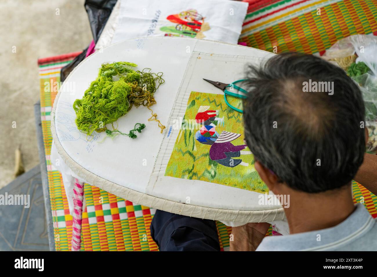 Ein vietnamesischer Handwerker wird von hinten gesehen, der eine farbenfrohe kulturelle Darstellung auf ein Tuch aufnäht. Das Bild unterstreicht das akribische Stockfoto