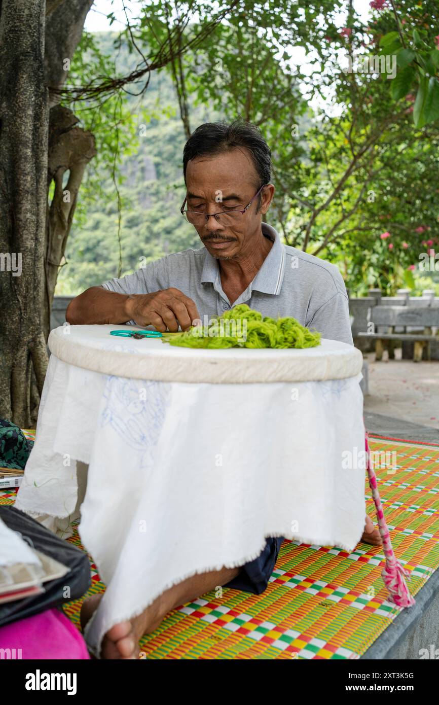 Ein älterer vietnamesischer Mann sitzt im Freien und näht sich akribisch auf einem runden Rahmen, der mit Stoffbäumen bedeckt ist, und die natürliche Kulisse verbessert sich Stockfoto