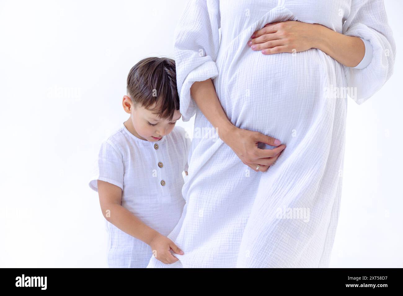 Nahaufnahme von Magen und Armen einer jungen schwangeren Frau in weißem Kleid und ihrem kleinen Sohn. Ein süßes Baby umarmt sein Mom-Bein. Die sanfte Berührung eines ch Stockfoto