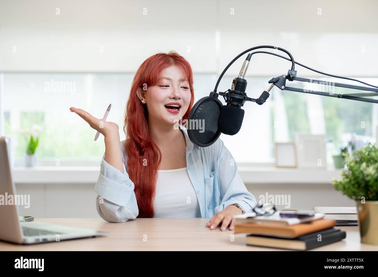 Eine charmante und fröhliche asiatische Podcaster mit rotem lockigem Haar spricht in ein Mikrofon und genießt die Aufnahme ihres Podcasts im Stockfoto
