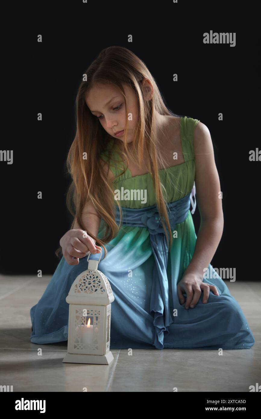 Niedliches kleines Mädchen mit langen Haaren und blauem Kleid, das auf dem Boden mit Laterne in gedämpfter Beleuchtung sitzt Stockfoto