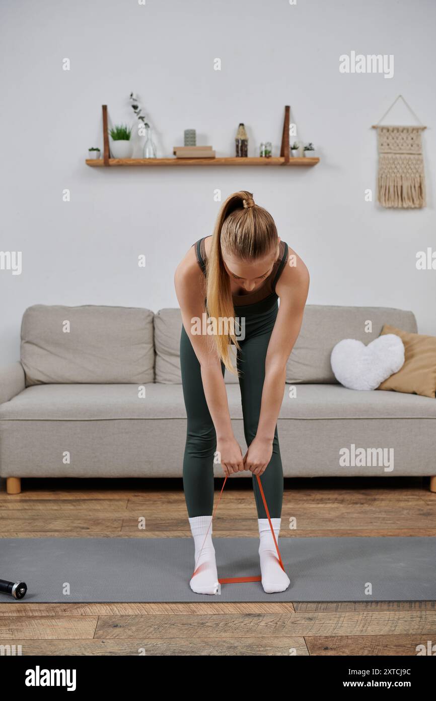 Frau in Workout-Kleidung dehnt sich mit einem roten Widerstandsband auf einer Yogamatte im Wohnzimmer. Stockfoto