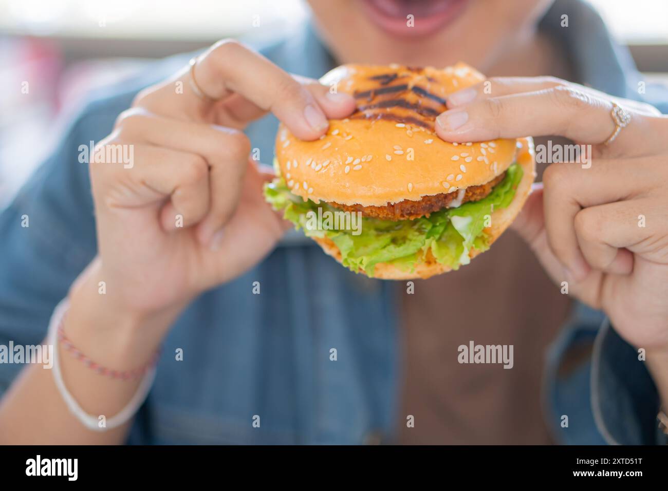 Eine Nahaufnahme einer Hand, die einen Sesambrötchen-Burger hält, gefüllt mit knusprigem gebratenem Huhn, frischem Salat und Mayonnaise, vor einem verschwommenen Hintergrund Stockfoto