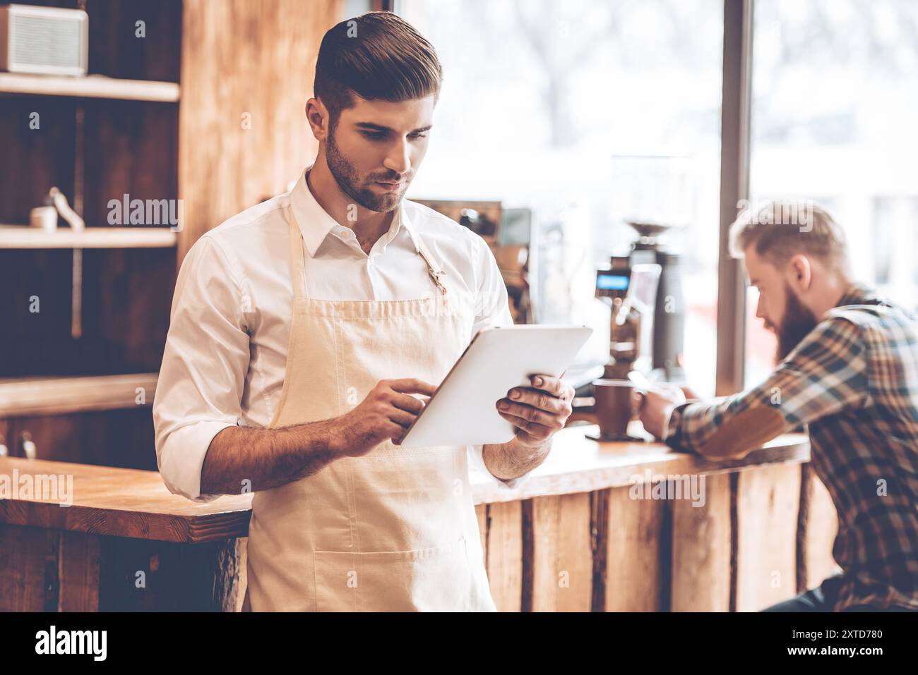 Neue Lösungen für sein Unternehmen. Junger, gutaussehender Mann in Schürze, der sein digitales Tablet benutzt, während er im Café mit einem Kunden im Hintergrund steht Stockfoto