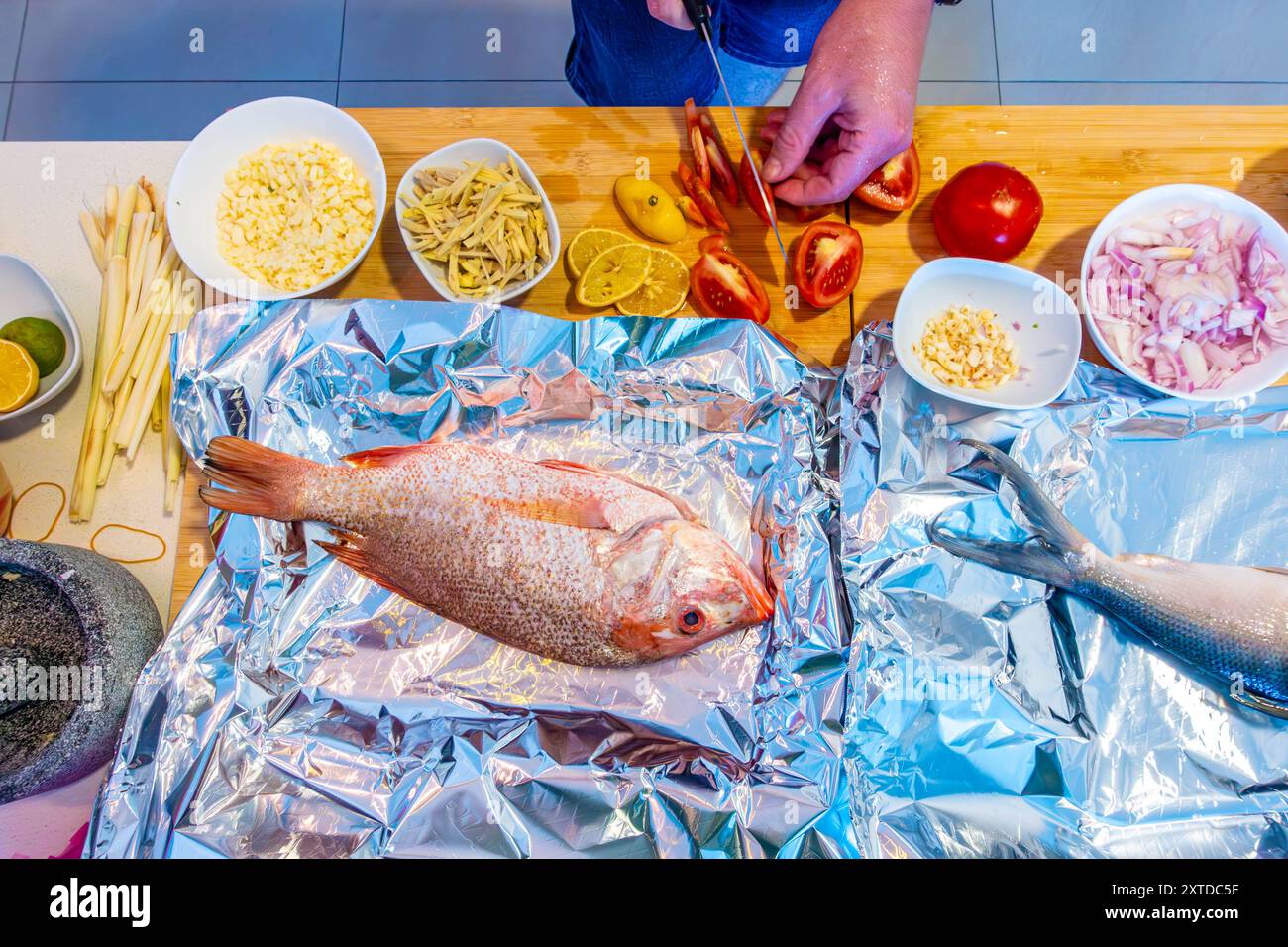 Wenn ein Mann Tomaten in Scheiben schneidet, um eine Mahlzeit mit rotem Schnapper, Gemüse und anderen Schüsseln gehackter Zutaten zuzubereiten. Stockfoto