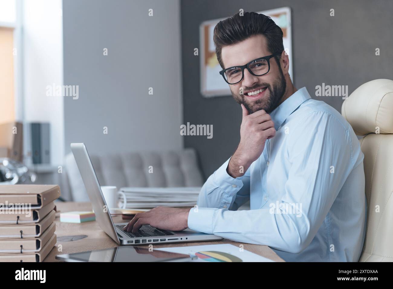 Selbstbewusst und erfolgreich. Seitenansicht eines hübschen jungen Mannes, der eine Brille trägt und mit einem Laptop arbeitet und mit einem Lächeln in die Kamera blickt, während er an seinem Arbeitsplatz sitzt Stockfoto