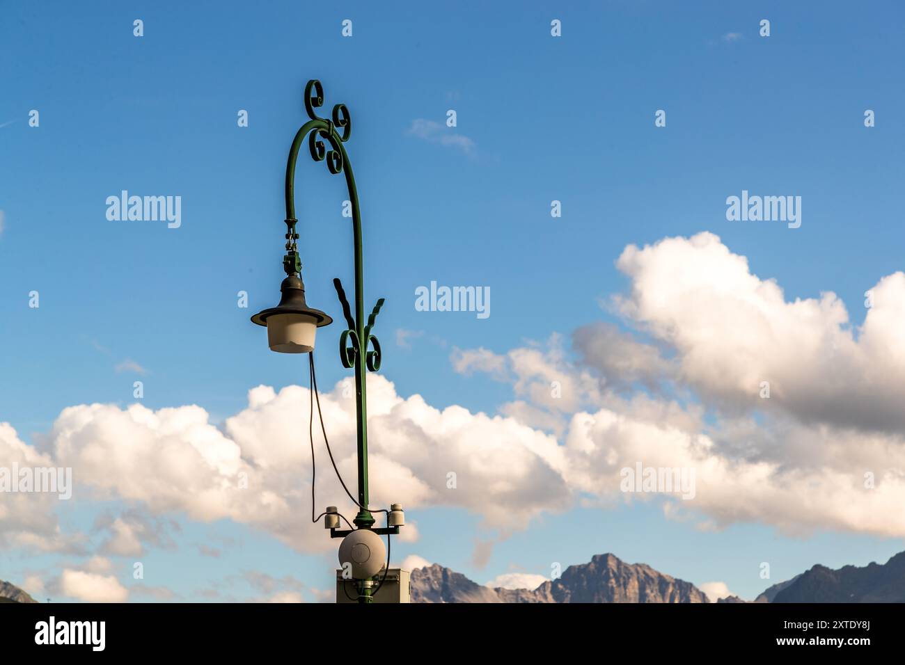 Eine schmiedeeiserne Gaslaterne mit geschwungenen Voluten erhebt sich vor Wolken und einem Gebirgskamm. Thomas-Mann-Weg, Davos, Graubünden, Schweiz. Stockfoto