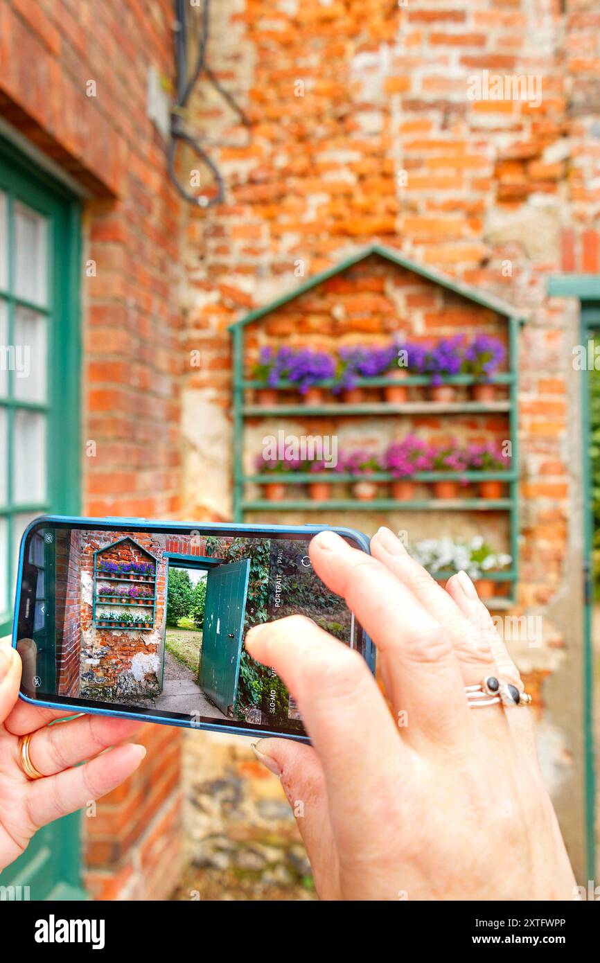 Hände halten ein iphone-Smartphone und machen ein Foto von einer rustikalen Ziegelwand mit grüner Tür und Blumenregal. Stockfoto