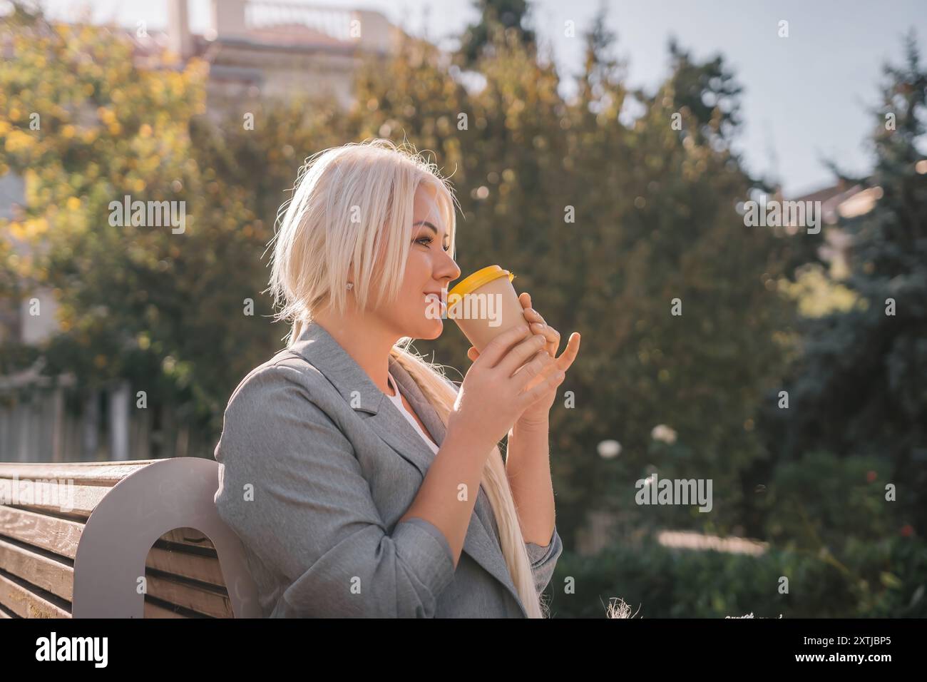 Eine blonde Frau sitzt auf einer Bank und trinkt Kaffee aus einer gelben Tasse. Sie trägt eine graue Jacke und hat ihr Haar im Pferdeschwanz. Die Szene ist friedlich Stockfoto